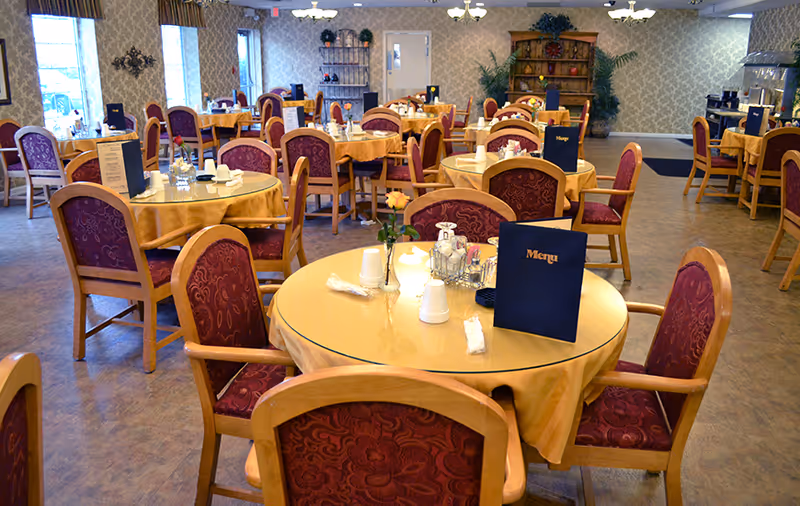 A spacious dining room with multiple round tables covered with yellow tablecloths and glass tops. Each table is surrounded by wooden chairs with red patterned cushions. Menus, napkins, cups, and condiments are placed on the tables. The room has patterned wallpaper, chandeliers, and a wooden hutch with decorative items in the background.