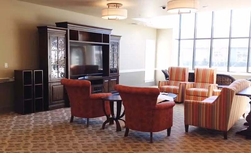 A cozy living room area with a large wooden entertainment center holding a flat-screen TV, two red upholstered armchairs, and a set of striped armchairs and a sofa arranged around a small round table. Large windows allow natural light to fill the room.