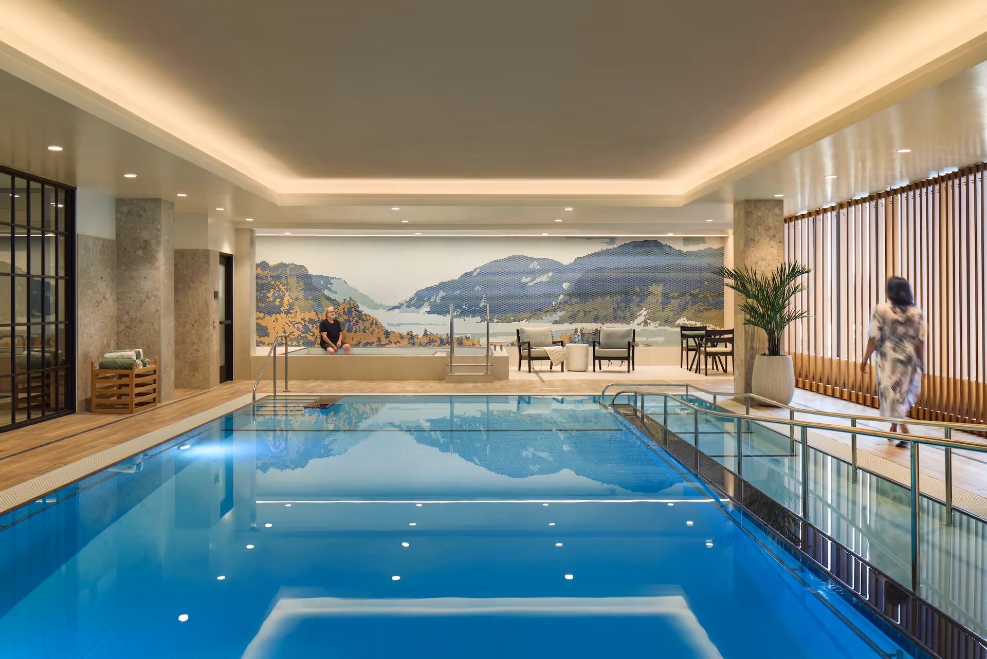 Indoor swimming pool area with clear blue water, surrounded by a wooden deck and glass railings. A person is sitting on the edge of the pool near a mural of a mountain landscape on the far wall. Another person is walking along the right side near large vertical wooden blinds and a tall potted plant. The ceiling has recessed lighting and a modern, clean design.