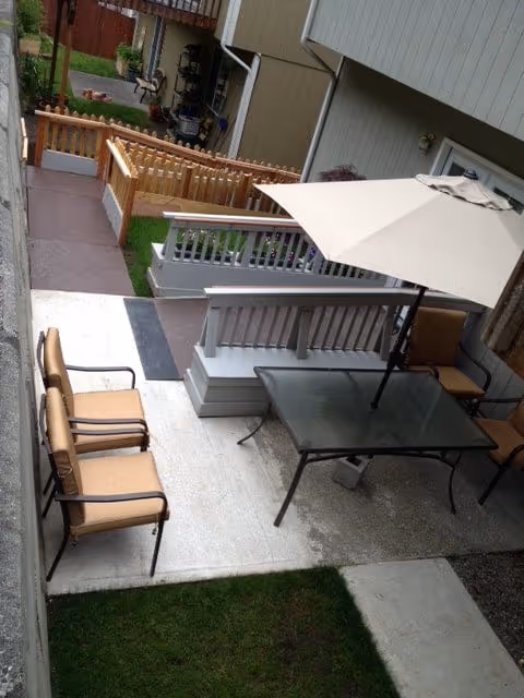 Backyard patio with a glass table and umbrella, several cushioned chairs, wooden railings, walkways and a small patch of grass between adjacent buildings.