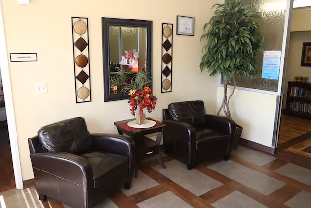 A waiting area with two dark brown leather armchairs separated by a small wooden table holding a vase with red and orange flowers. A large mirror is mounted on the wall above the table, flanked by two decorative wall hangings. To the left, a door labeled 'Admissions' is partially visible. A tall green plant is placed in the corner near a frosted glass window with a blue notice attached. The floor has a patterned design with wood and tile elements.
