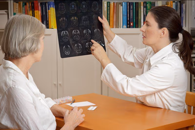 A female doctor in a white coat is showing and explaining brain scan images to an elderly woman seated at a wooden table in a room with bookshelves in the background.