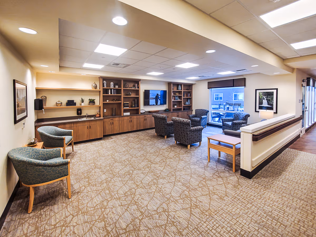 A spacious and well-lit common area in Marquis Eugene Assisted Living featuring comfortable armchairs arranged around a coffee table, built-in wooden shelves and cabinets along the wall with decorative items, a flat-screen TV mounted on the wall, and a large window showing an outdoor patio with tables and chairs.