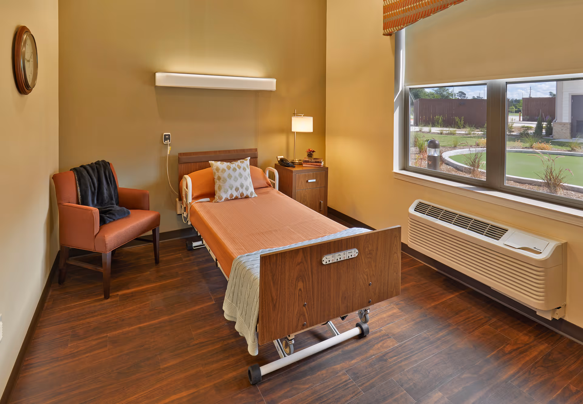 A single-occupancy senior care bedroom with a hospital-style bed, armchair, nightstand and a large window overlooking landscaped grounds.
