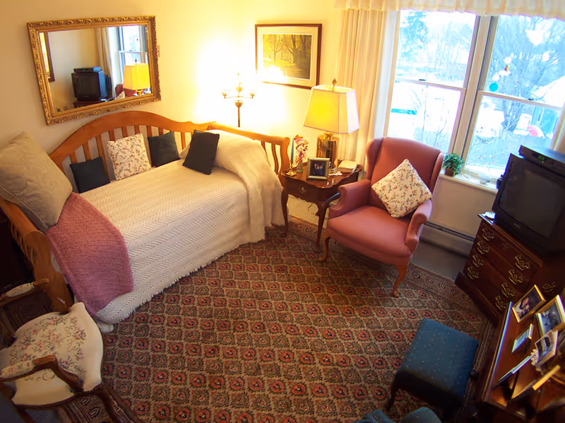 Cozy living room with a wooden daybed covered with a white knitted blanket and several pillows, a pink armchair with a floral pillow, a wooden side table with a lamp and framed photos, a large window with curtains, a patterned carpet, a wooden dresser with an old-style TV, and a large mirror on the wall reflecting part of the room.