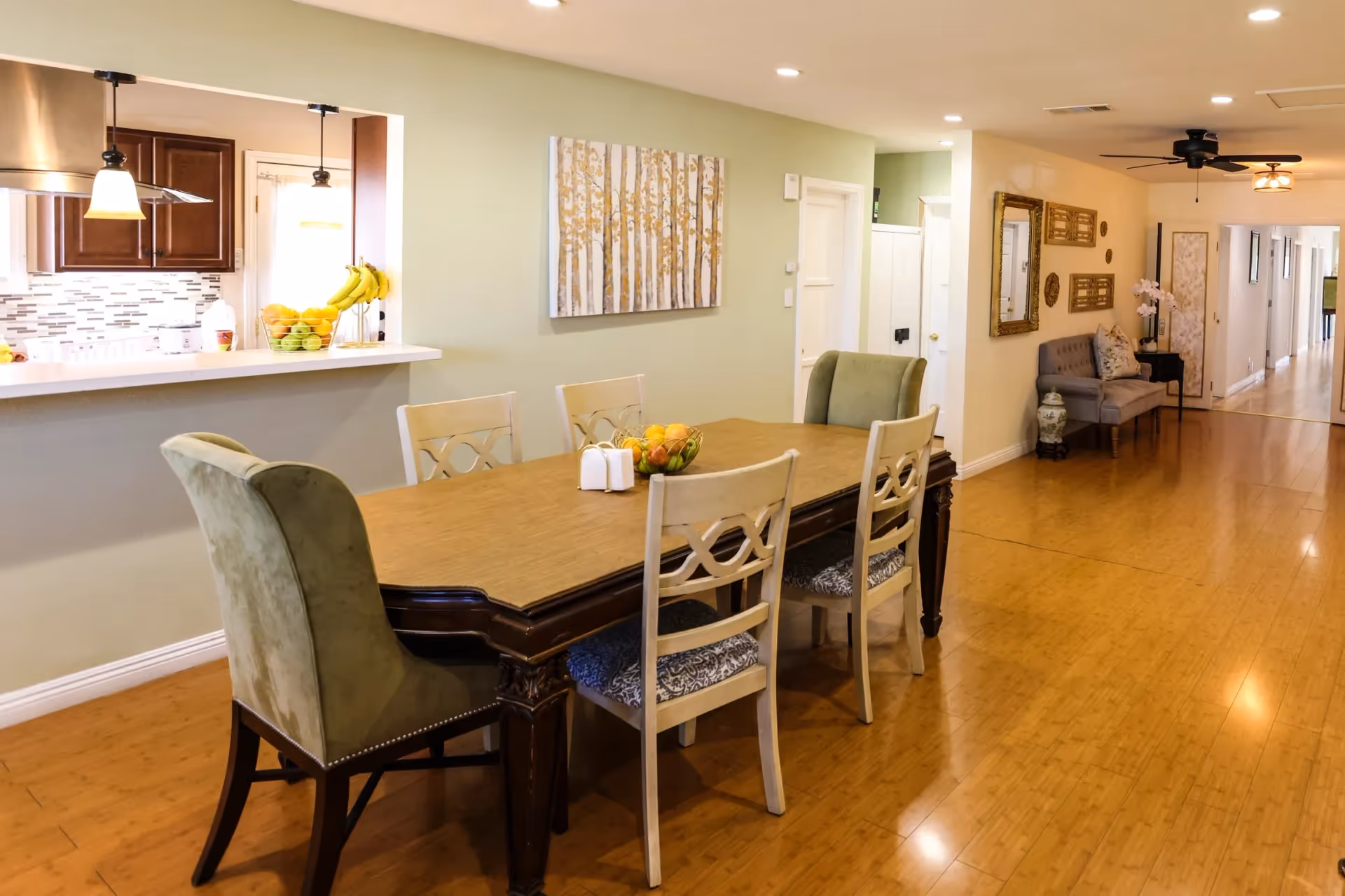 Dining area with a wooden table and mixed chairs beside an open kitchen counter in a bright senior living interior.