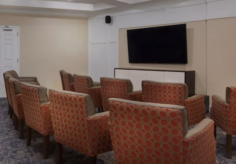 A small theater-style room with two rows of red patterned armchairs facing a large flat-screen TV mounted on a beige wall. The room has a carpeted floor and a white door on the left side.
