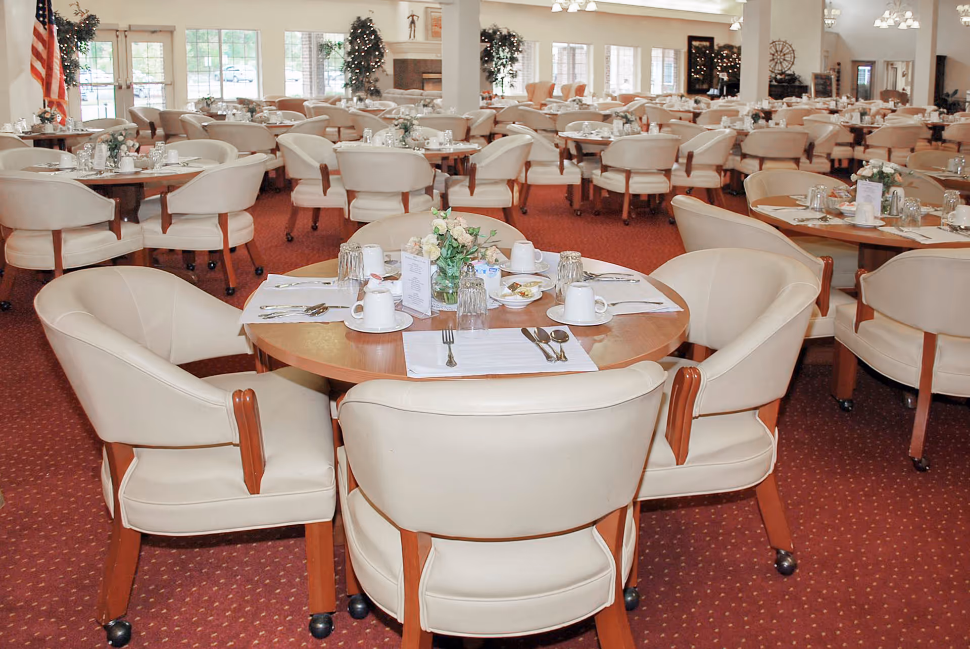 Spacious dining room with round wooden tables set for meals and cream upholstered chairs on a red carpet.