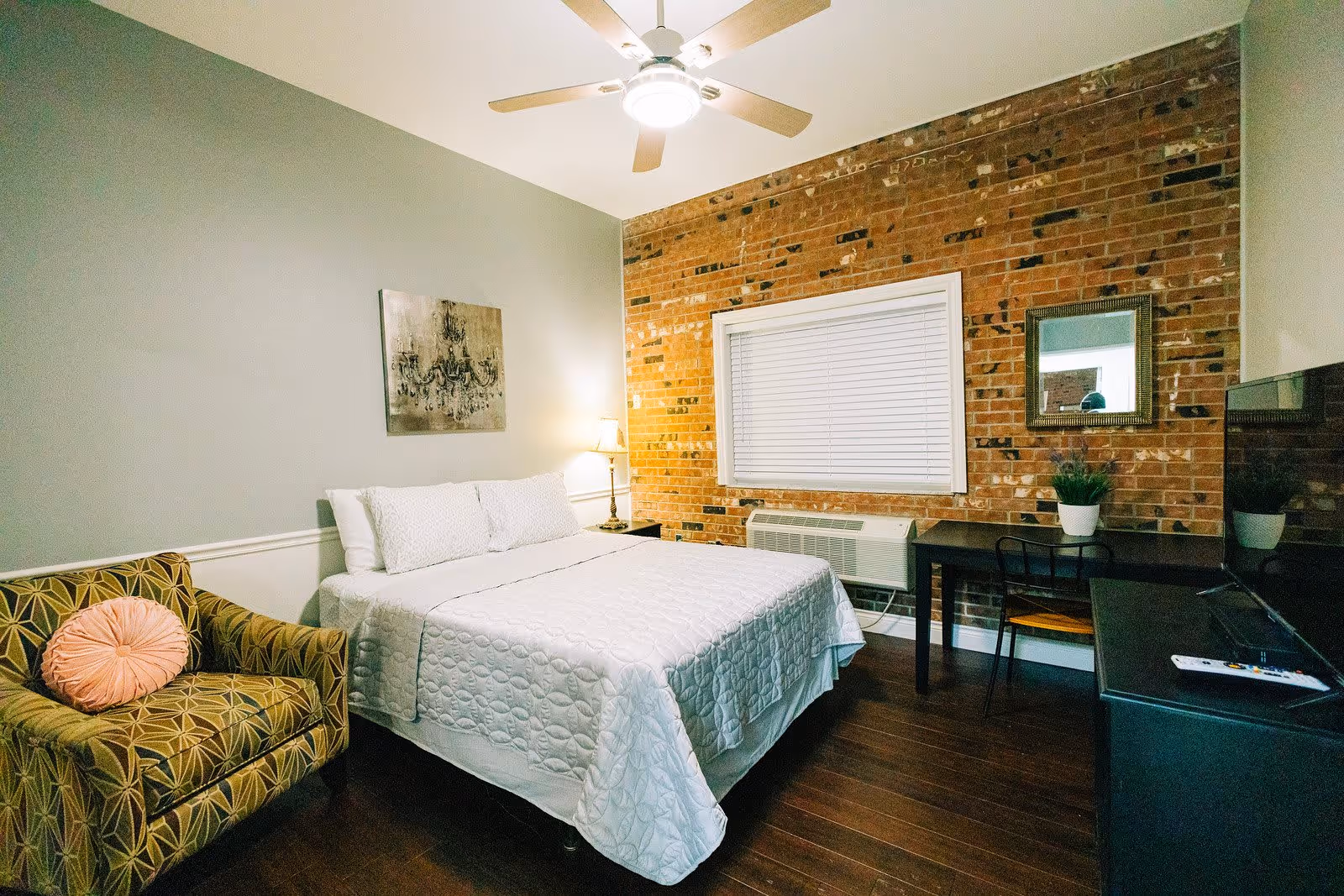 A cozy bedroom with a white bedspread on a double bed, a patterned armchair with a round pink pillow, a brick accent wall with a window covered by white blinds, a desk with a chair and a potted plant, a wall mirror, a TV on a stand, and a ceiling fan with lights.