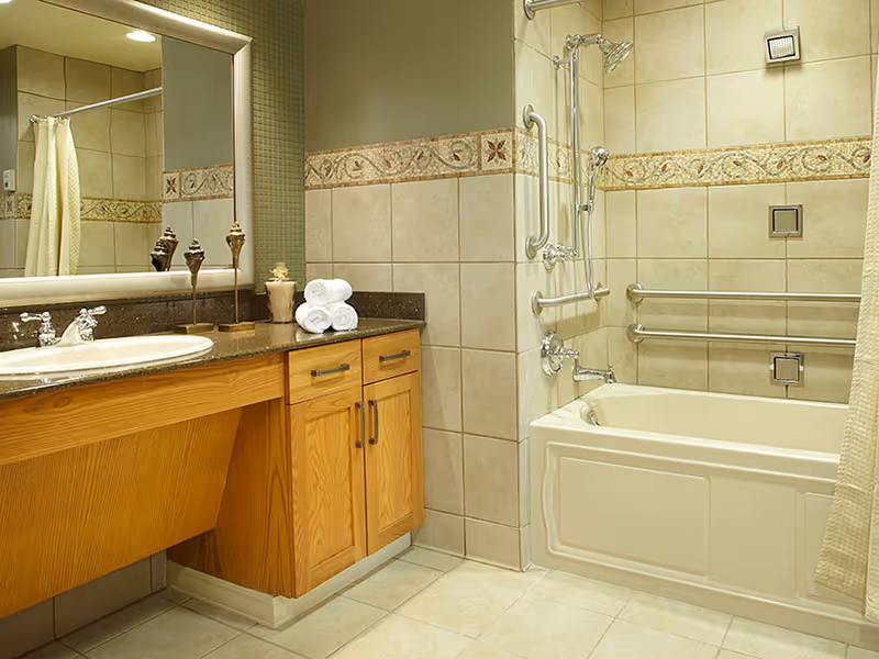 A bathroom with a wooden vanity featuring a sink and countertop, a large mirror above it, and a bathtub with grab bars and a showerhead. The walls are tiled with decorative trim, and there is a shower curtain partially visible.