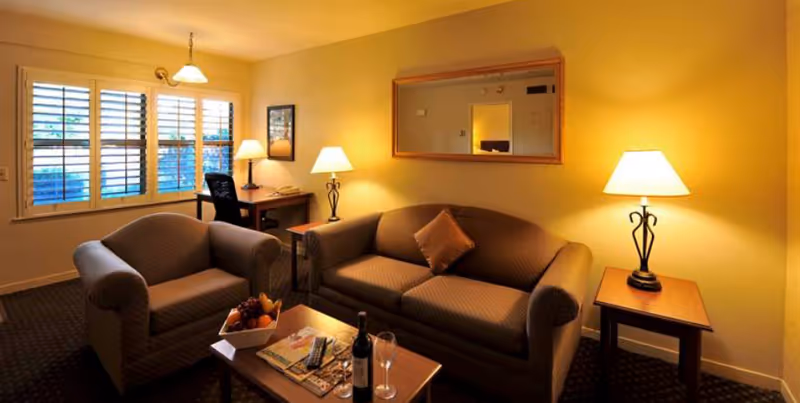 A cozy living room with a brown sofa and matching armchair, a wooden coffee table with a bottle of wine, two wine glasses, a fruit bowl, and magazines. There are two side tables each with a lamp, a desk with a chair near a window with white shutters, and a large mirror on the wall reflecting part of the room.