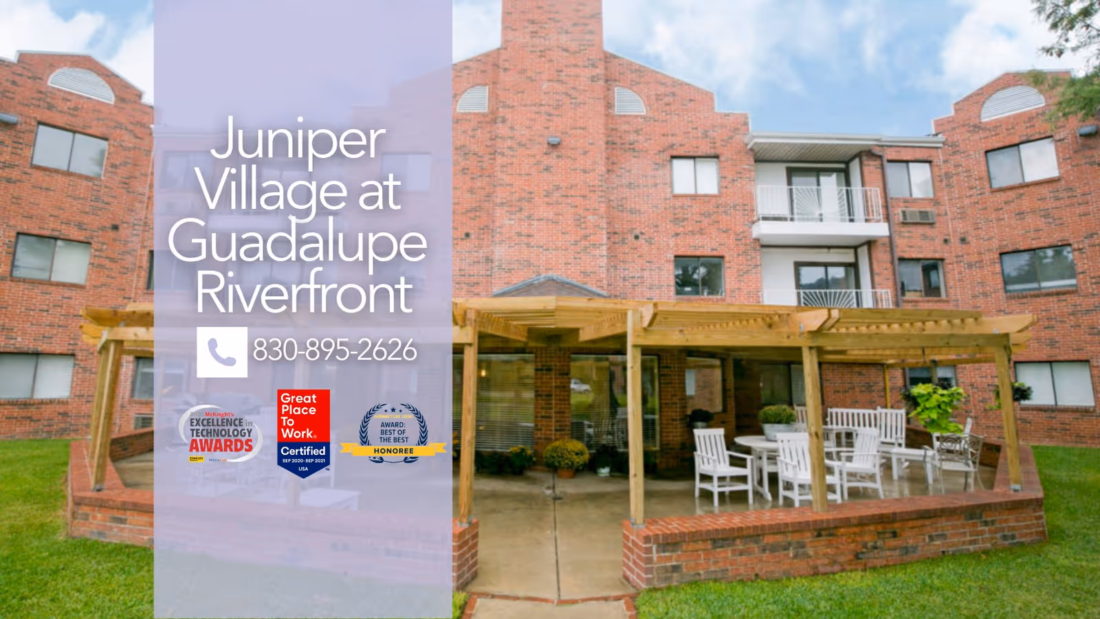 Exterior view of a brick senior living facility building with multiple windows and balconies. In front of the building is a covered patio area with a wooden pergola, white outdoor chairs, and tables on a concrete surface surrounded by grass.