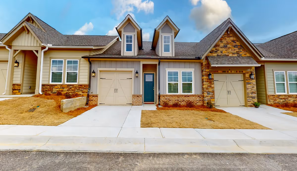 Front exterior view of a senior living facility unit at The Lodge at Stephens Lake, featuring beige siding, stone accents, two garage doors, a green front door, and two dormer windows on the roof under a partly cloudy sky.