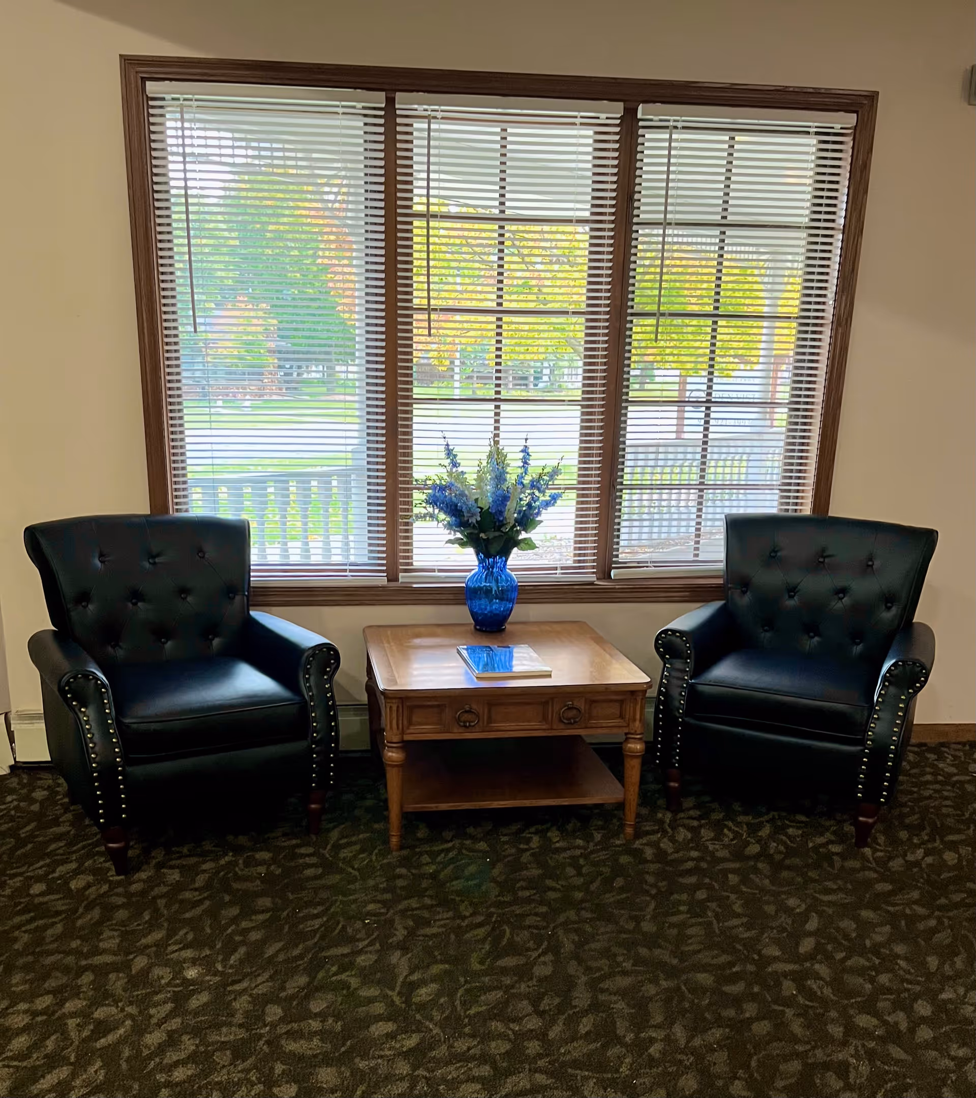 Two black leather armchairs with button tufting and nailhead trim flank a wooden side table with a blue vase of flowers and a book, set in front of a large window with wooden blinds showing a view of green trees outside.