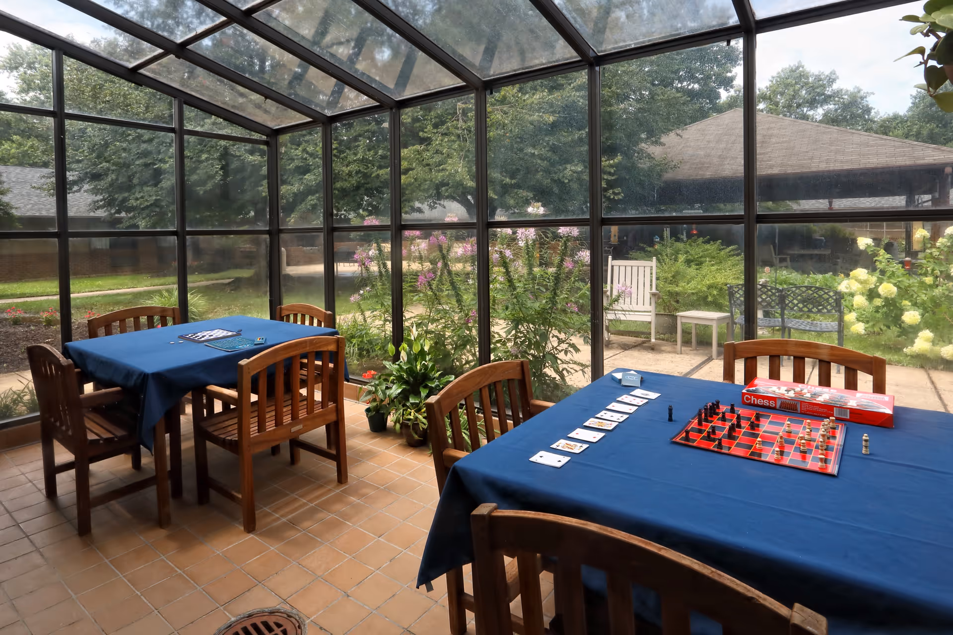 Sunroom-style common room with wooden tables and chairs covered in blue tablecloths, a chess set and playing cards on the tables, and large glass windows looking out to a garden.