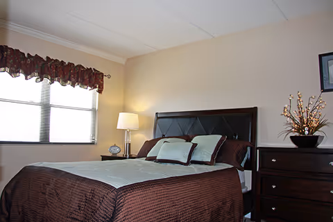 A neatly made bedroom with a bed, nightstand lamp, dresser, and a window with blinds.