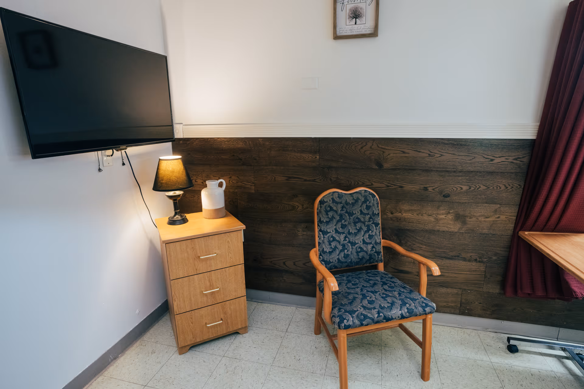 A corner of a room with a wooden side table holding a lamp and a white pitcher, a blue patterned upholstered wooden chair, a wall-mounted flat screen TV, a wooden panel on the wall, a framed picture above, and a maroon curtain partially visible on the right.