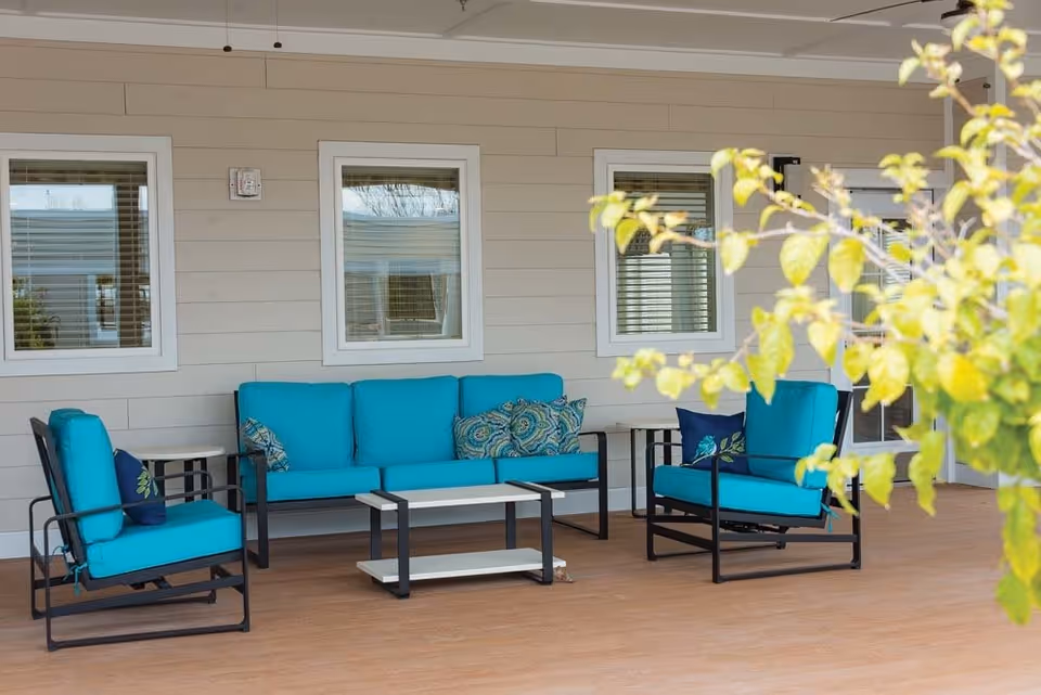 Outdoor patio area with turquoise cushioned seating including a sofa and two armchairs, a coffee table, and side tables. The background shows a beige wall with three windows and a door, and a leafy plant partially visible on the right side.