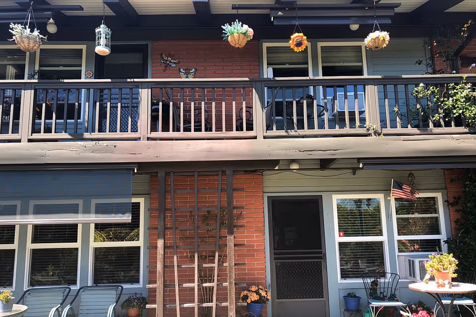 Front exterior of a two-story residential building with a balcony, hanging plants, patio chairs, potted flowers, and an American flag.