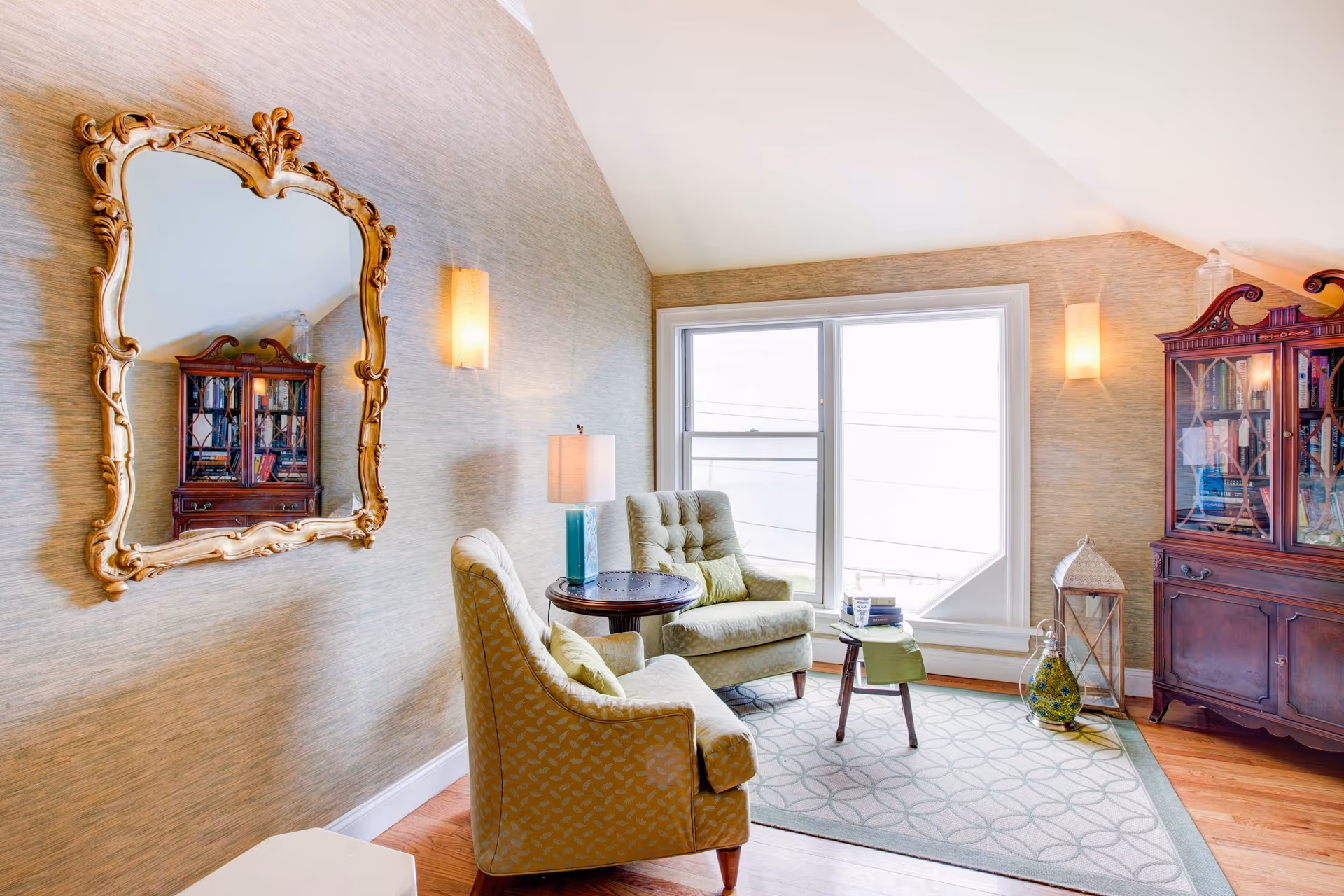 A cozy sitting area with two upholstered armchairs and a small round table with a lamp between them. The room features a large window letting in natural light, a decorative gold-framed mirror on the wall, a patterned area rug, and a dark wooden cabinet with glass doors displaying books and decorative items.