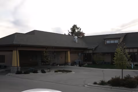 Exterior view of a single-story assisted living facility building with a large roof, several windows, small trees, and a paved driveway in front under a cloudy sky.