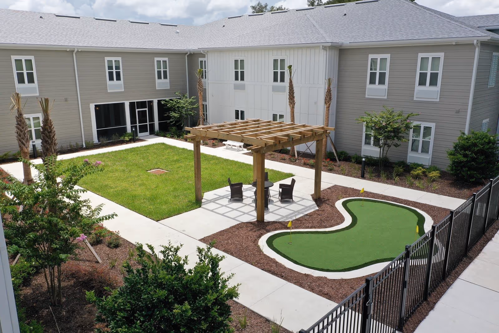 Outdoor courtyard area at The Canopy at Walden Woods featuring a green lawn, a wooden pergola with three chairs and a small table underneath, a putting green with three yellow flags, surrounded by a paved walkway and landscaping with trees and shrubs, enclosed by a black metal fence.