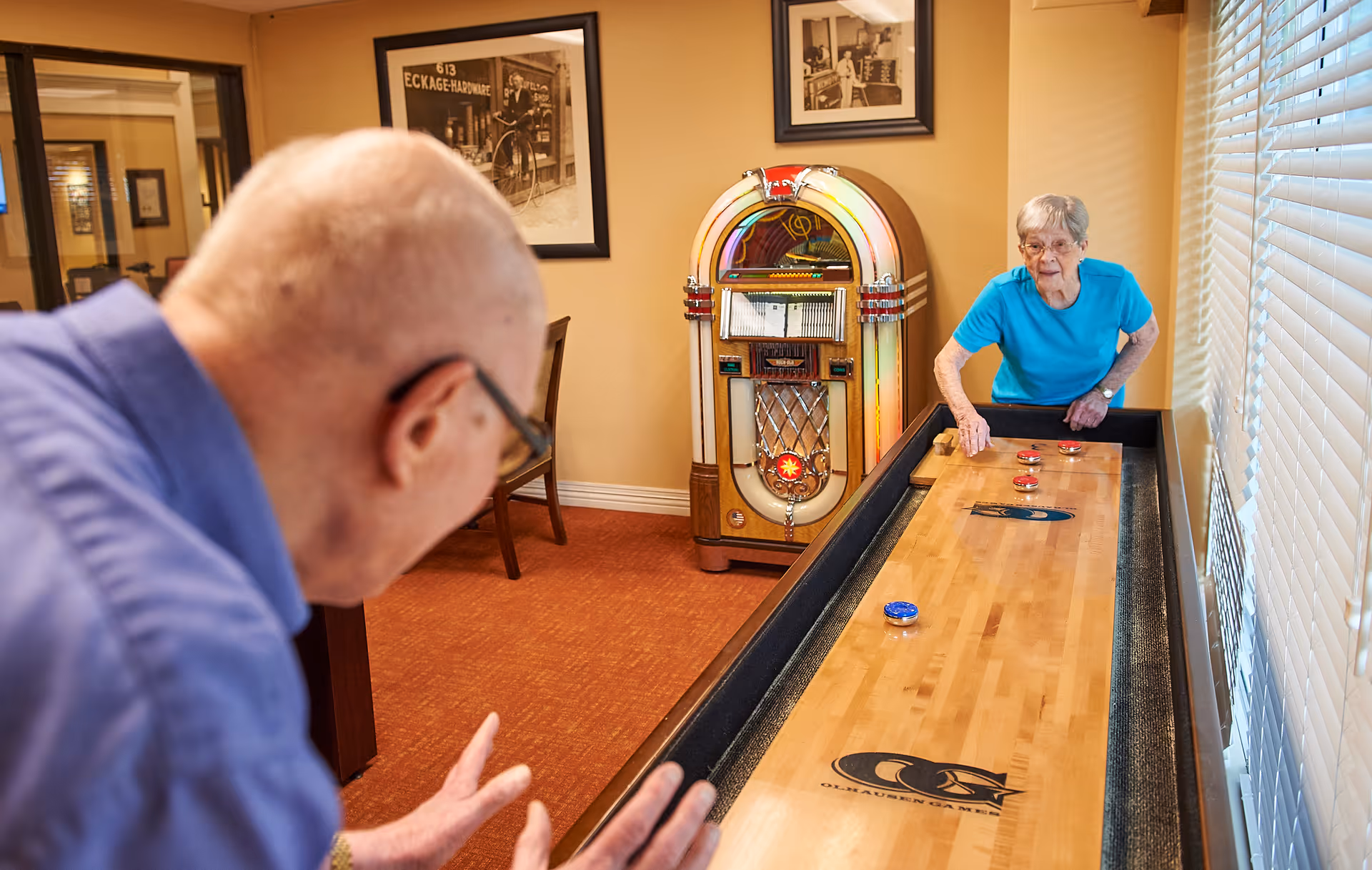 An elderly man and woman playing shuffleboard indoors in a room with warm lighting. The woman is wearing a blue shirt and glasses, standing near the shuffleboard table, while the man is in the foreground preparing to play. Behind them is a vintage jukebox and framed black-and-white photos on the wall.