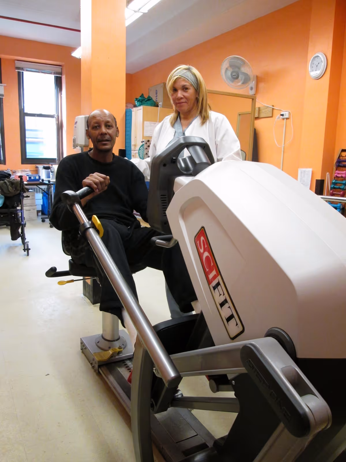 A man is seated on a SciFit exercise machine in a room with orange walls, accompanied by a woman in a white coat standing beside him. The room appears to be a rehabilitation or therapy area with various equipment and supplies in the background.