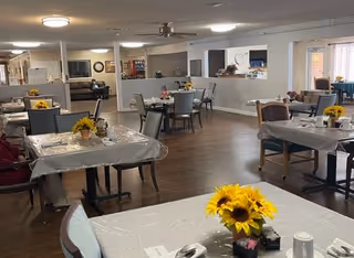 A spacious dining area in a senior living facility with multiple tables covered in plastic tablecloths, each decorated with a vase of sunflowers. Chairs are arranged around the tables, and the room has wood flooring and neutral-colored walls. In the background, there is a living area with sofas and a television.