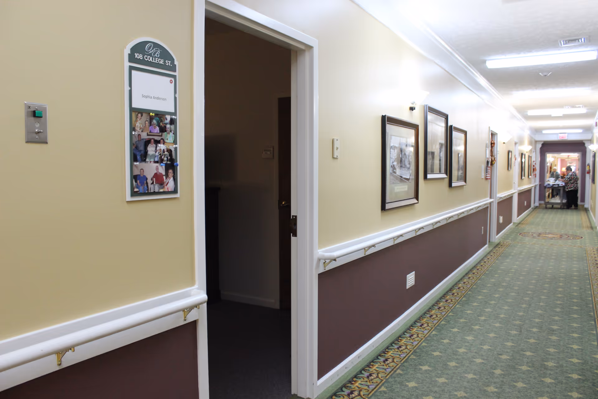A long hallway inside a senior living facility with beige and brown walls, green patterned carpet, handrails along the walls, framed pictures hanging on the right wall, and a partially open door on the left. At the end of the hallway, two people are seen near a cart.