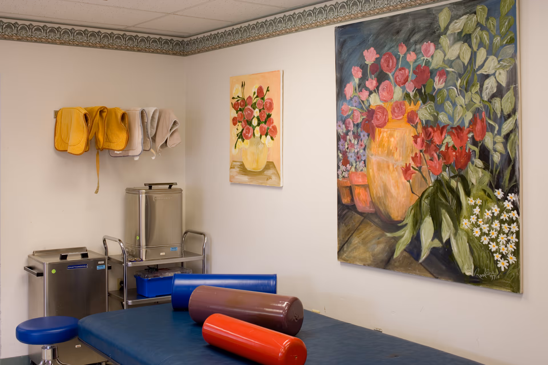 A therapy or treatment room with a blue padded table and cylindrical bolsters in red and brown. The walls are decorated with two paintings of flower arrangements in vases. There are folded towels hanging on the wall and stainless steel medical equipment carts in the corner.