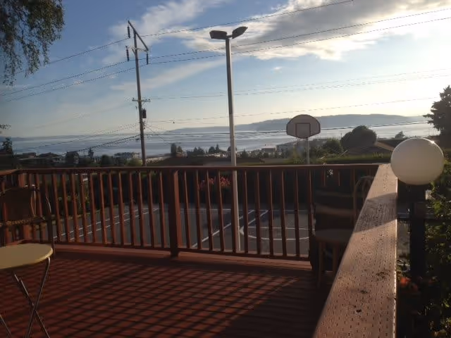 Wooden deck with railing and chairs overlooking a parking lot, basketball hoop and waterfront view.