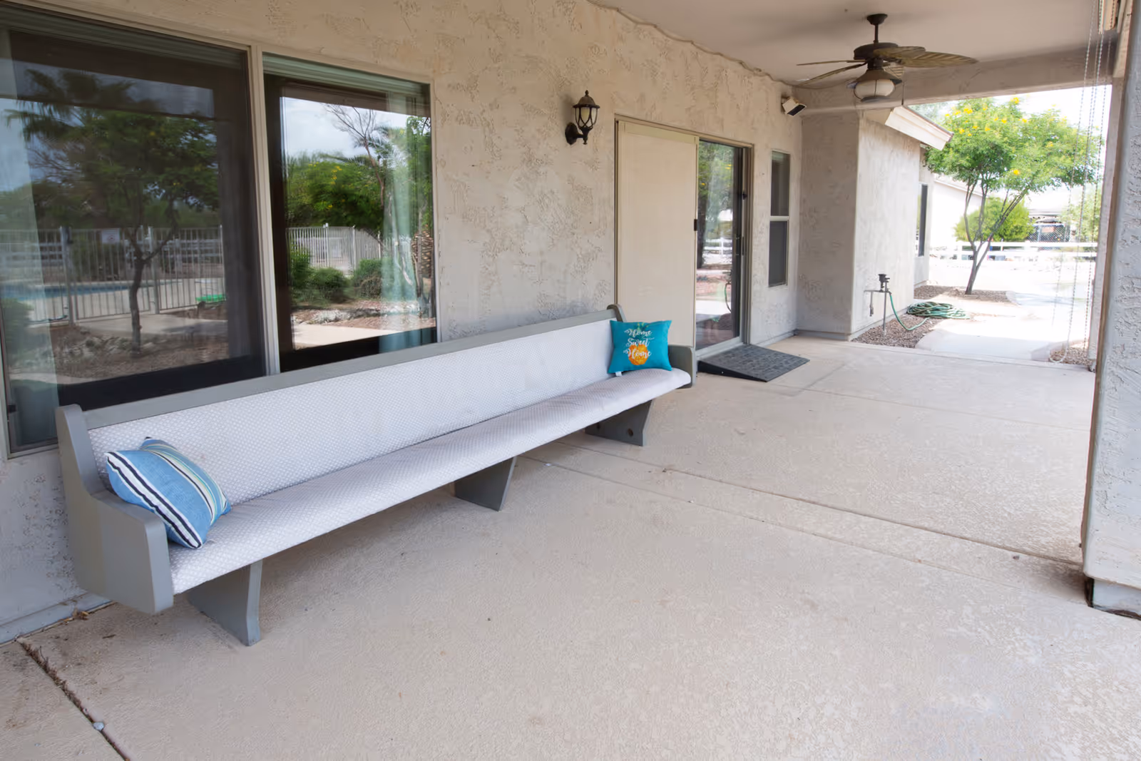 Covered exterior patio with a long cushioned bench and throw pillows, sliding glass doors, a ceiling fan, and a view of a fenced yard.