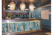 Reception area of a care center with a front desk featuring a decorative panel with blue and green patterns. Three pendant lights hang from the ceiling above the desk. The walls are painted blue, and there are shelves and a door visible in the background.