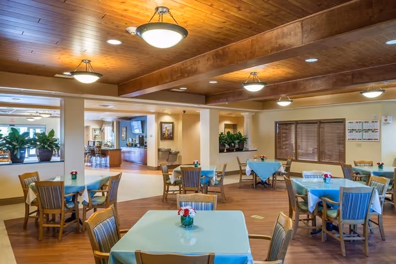 A bright dining room with multiple tables covered in light blue tablecloths, each adorned with a small vase of red and white flowers. Wooden chairs surround the tables, and the room features a wooden ceiling with recessed lighting. Large windows with blinds and potted plants add to the welcoming atmosphere.