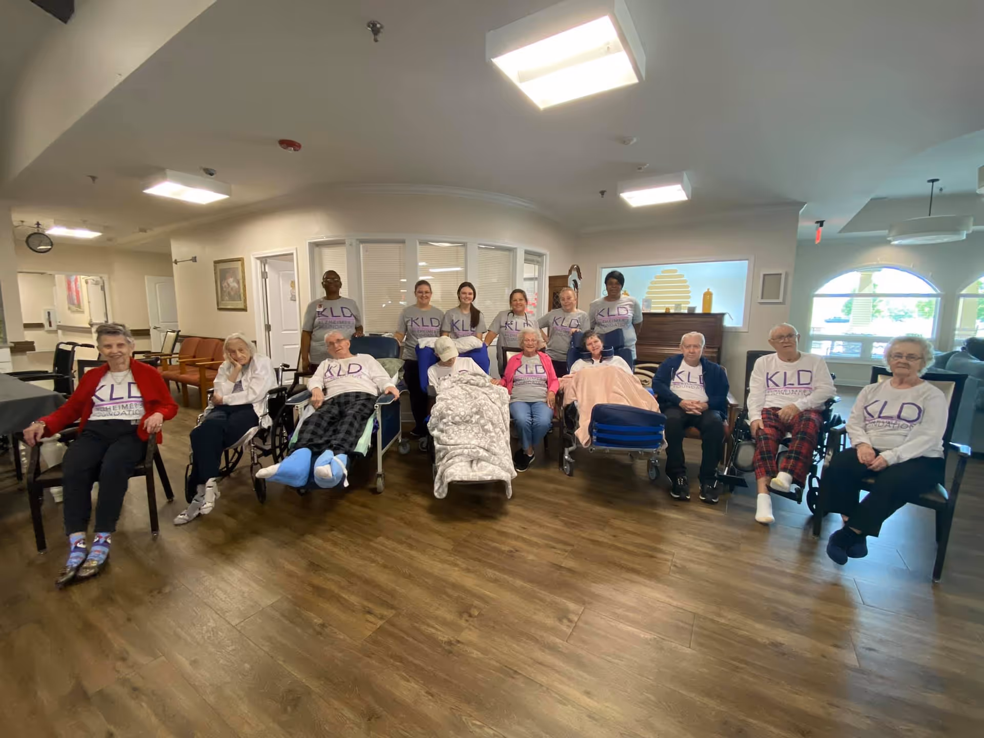 A group of elderly residents and staff wearing matching KLD shirts seated and in wheelchairs in a memory care facility common room.