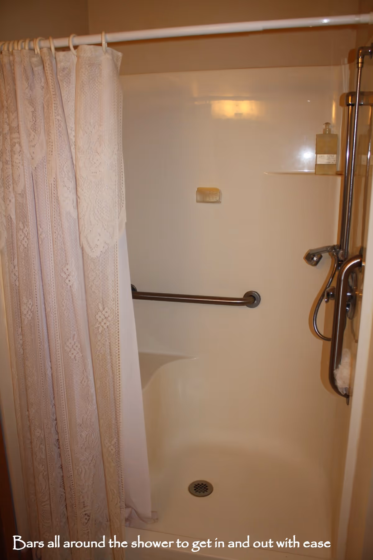 A shower area with a white lace shower curtain partially drawn to the side. Inside the shower, there is a horizontal grab bar on the back wall and a vertical grab bar near the shower controls. A soap dish and a bottle of liquid soap are visible on a small shelf. The shower floor has a drain in the center.