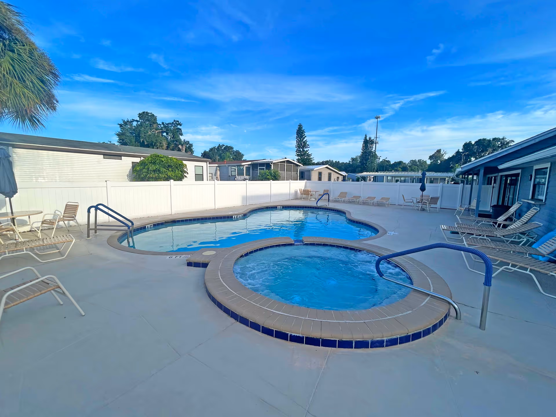 Outdoor pool and hot tub with lounge chairs on a fenced patio at a retirement community under a blue sky.