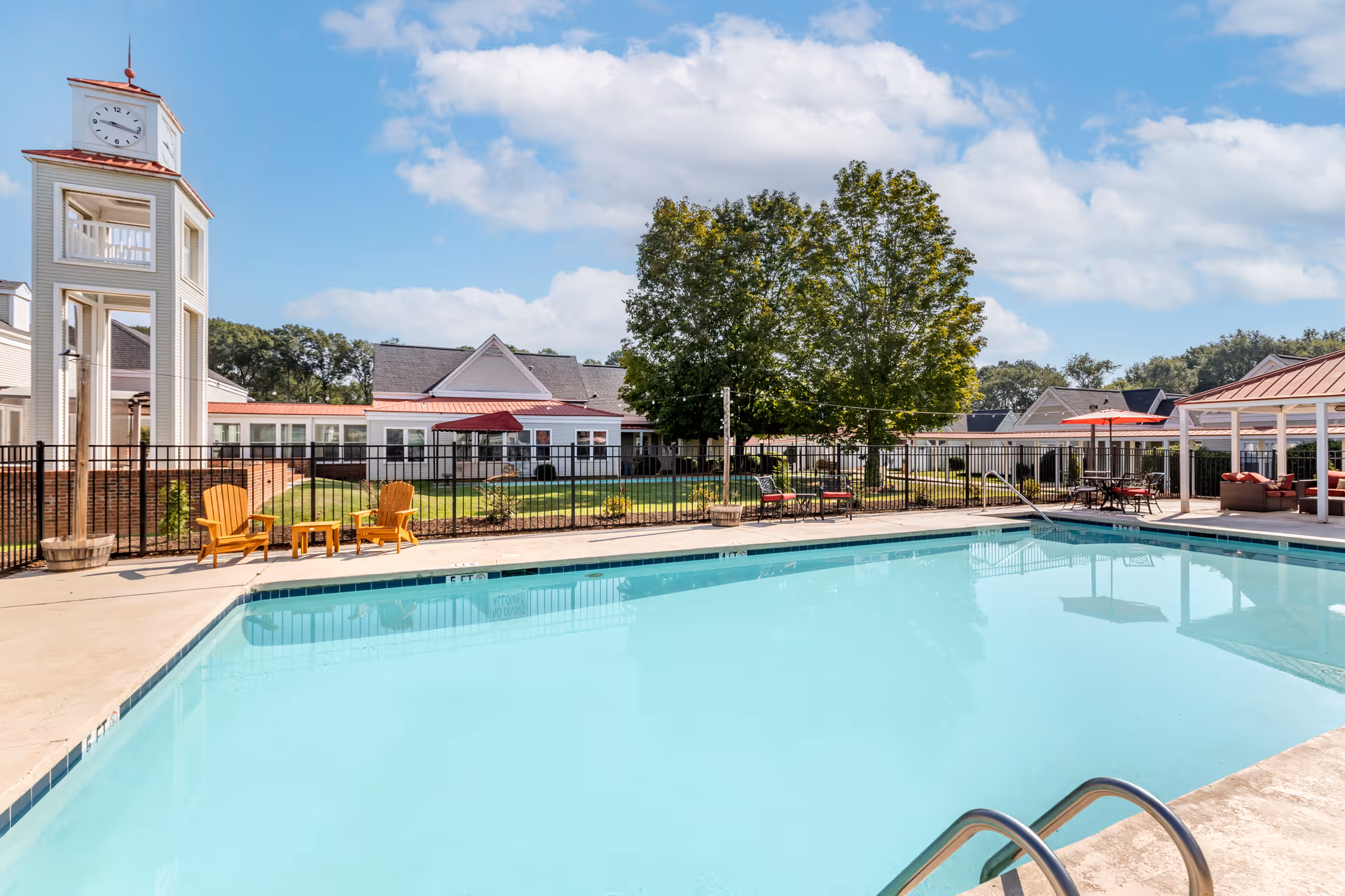 A clear outdoor swimming pool surrounded by deck chairs, umbrellas, a fence, and nearby residential buildings with a clock tower.