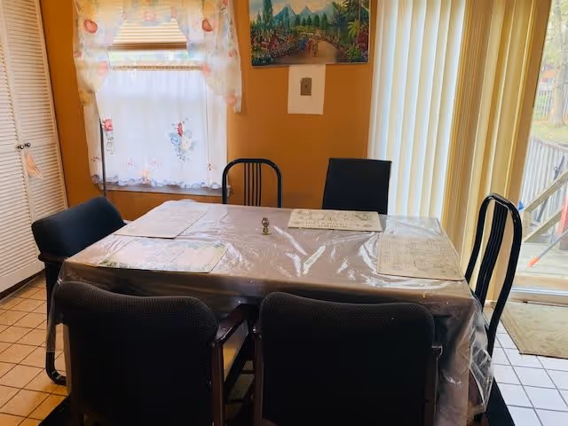 A dining area with a rectangular table covered by a clear plastic tablecloth and four black chairs around it. The table has placemats and a small decorative item in the center. There is a window with floral curtains on the left wall and vertical blinds covering a sliding glass door on the right side. The walls are painted a warm yellow color, and a colorful landscape painting hangs above the table.