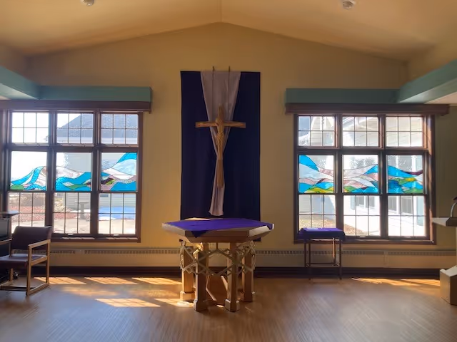 Interior view of a chapel or prayer room with a wooden crucifix mounted on a purple cloth backdrop, flanked by two large windows with colorful stained glass panels. There is a wooden altar covered with a purple cloth in the center and chairs on either side. Sunlight streams through the windows onto the wooden floor.