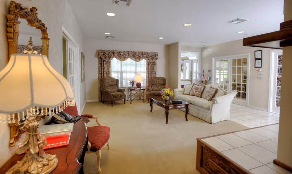 A cozy living room area with a beige carpet and tiled floor section. The room features a white sofa with floral cushions, two patterned armchairs by a window with floral curtains, a wooden coffee table with a vase of flowers, and a side table with a lamp. There is also a wooden console table with a decorative lamp and a red cushioned chair. The walls are light-colored, and there are glass-paneled doors leading to another room.