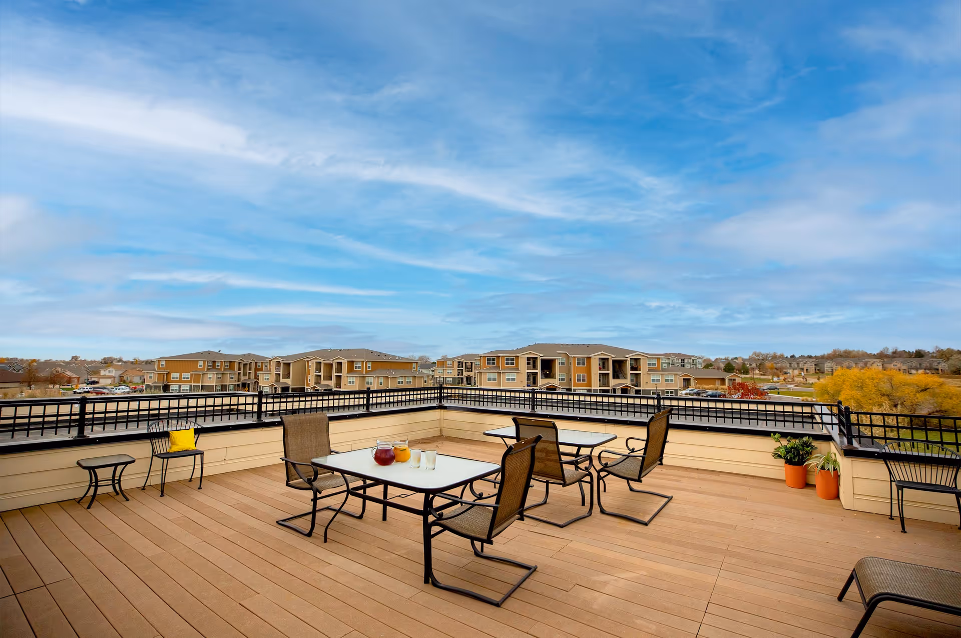 A spacious outdoor patio area with wooden flooring, featuring two tables with chairs around them. One table has a pitcher of red beverage, a pitcher of orange beverage, and two glasses. There are potted plants in the corner and a small side table with a yellow cushion on a chair. The patio is enclosed by a black metal railing and overlooks a residential neighborhood with multiple apartment buildings under a partly cloudy blue sky.