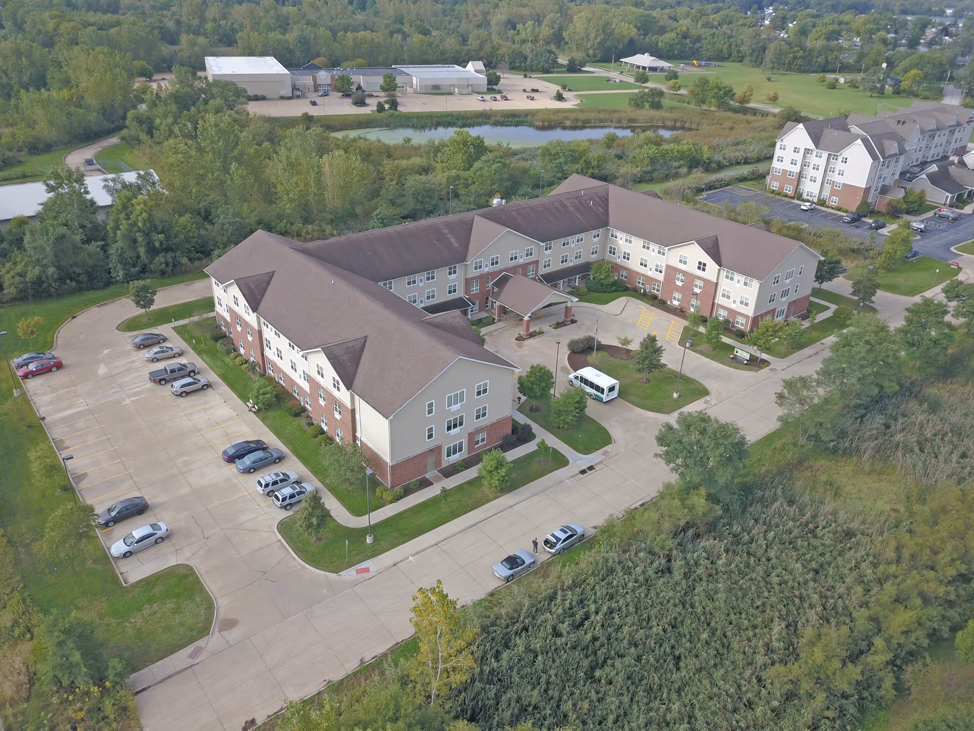 Aerial view of a three-story L-shaped senior living building with an entrance canopy, parking lot, surrounding trees and a nearby pond.