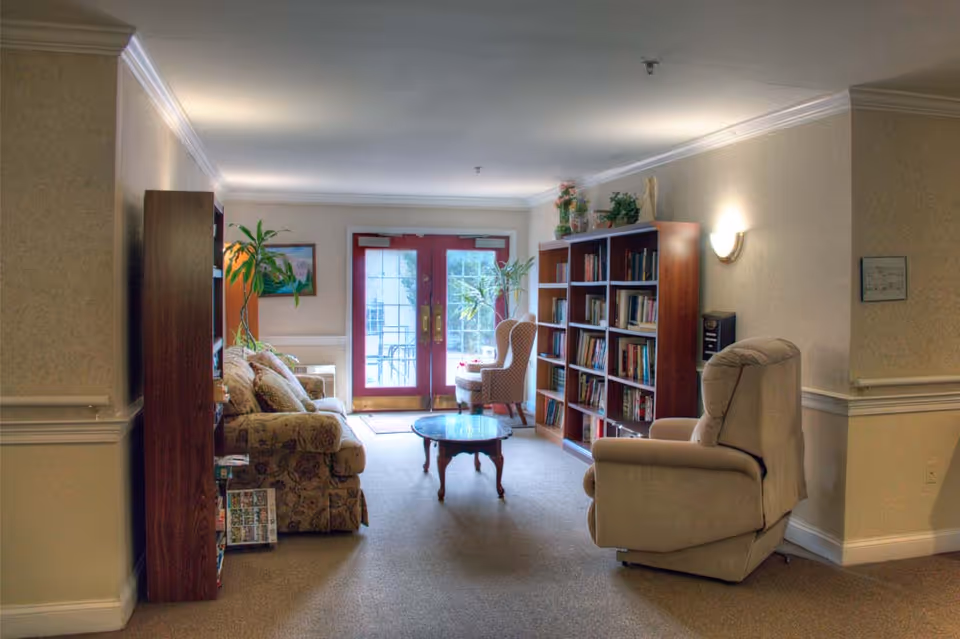 A cozy living room area in Merion Gardens Assisted Living featuring a floral patterned sofa, a beige recliner, a wooden coffee table, and bookshelves filled with books. There are two large glass doors at the far end letting in natural light, with a small table and chair near the doors. The walls are light-colored with crown molding and a wall sconce providing additional lighting.
