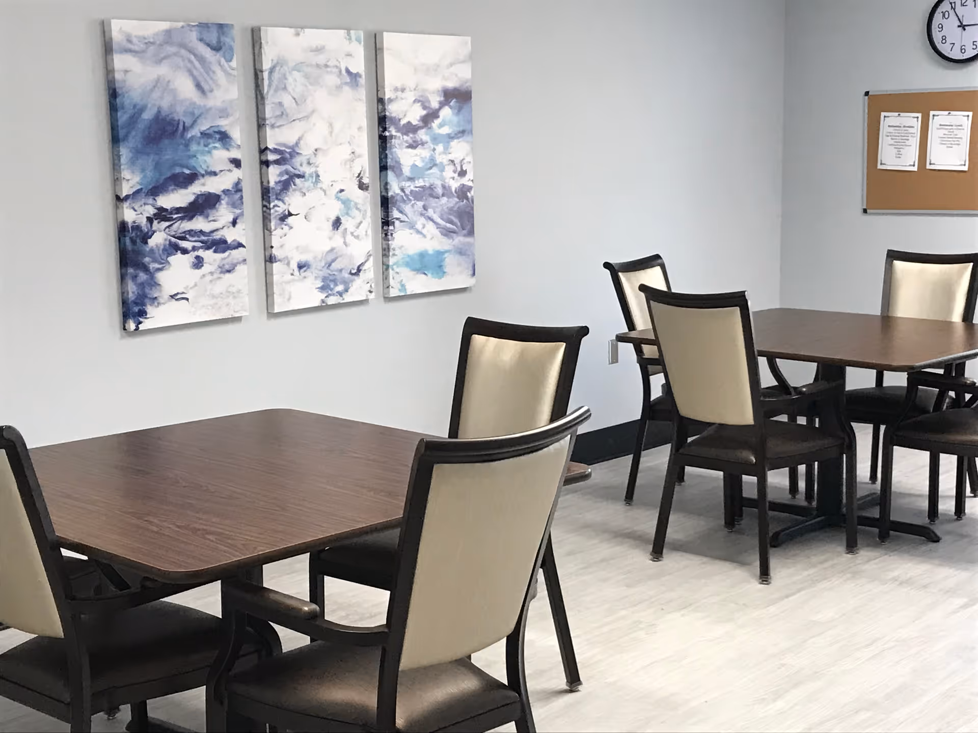 Dining room with several wooden tables and beige-upholstered chairs, a blue abstract triptych on the wall, and a bulletin board with a clock.