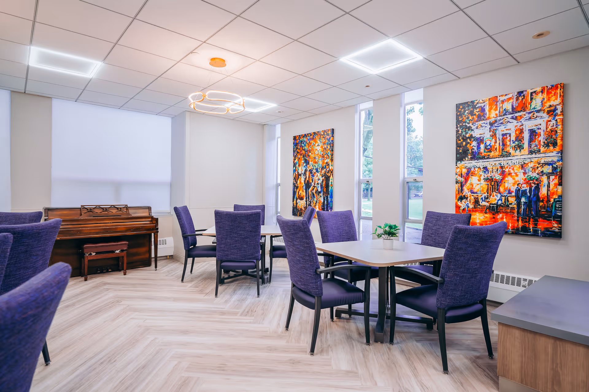 A bright and modern common area with several tables and purple upholstered chairs arranged around them. The room features large windows letting in natural light, colorful abstract paintings on the walls, a wooden piano with a bench, and a contemporary circular ceiling light fixture. The floor has a light wood pattern.