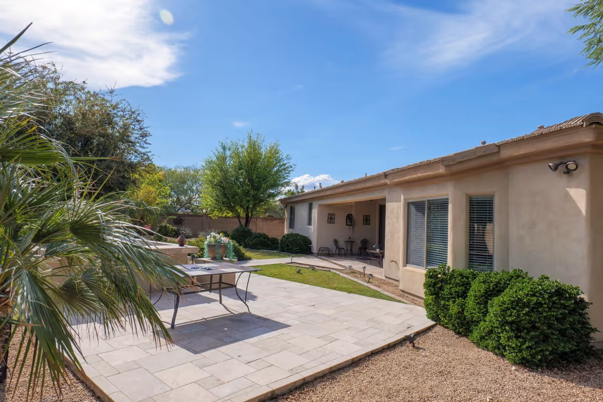 Outdoor patio area of a senior living facility with a tiled floor, a table, potted plants, green bushes, trees, and a beige building with windows and a covered seating area under a clear blue sky.