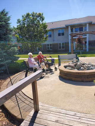Two elderly women sitting on benches near a circular stone fire pit in an outdoor courtyard area of a senior living community. One woman is using a walker. The background shows a two-story building with windows, trees, and a wooden pergola.