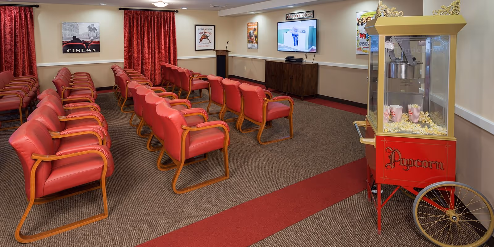 A small theater room with rows of red cushioned chairs with wooden armrests facing a wall-mounted TV screen. There is a red and gold popcorn machine filled with popcorn on the right side of the room. The walls are decorated with movie posters and red curtains cover the windows.